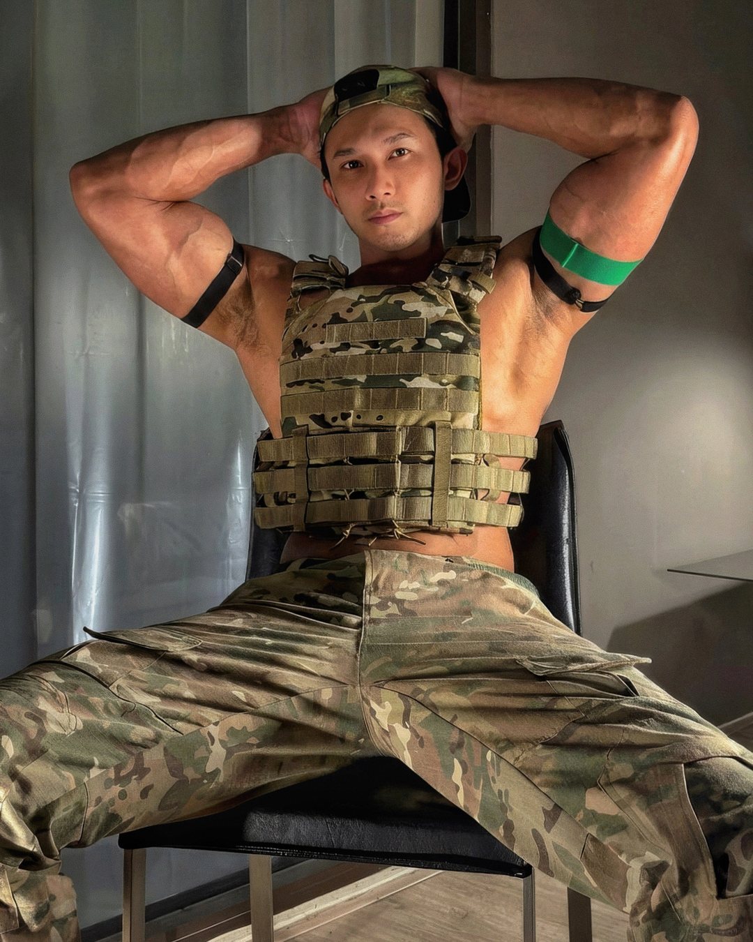 spicy somtam in a camo cap and tactical vest, hands behind his head, sitting with his legs apart