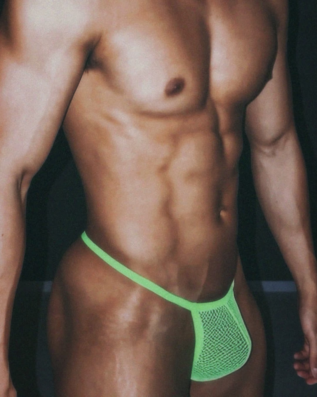 torso shot of a muscular man in a neon green mesh thong against a dark background